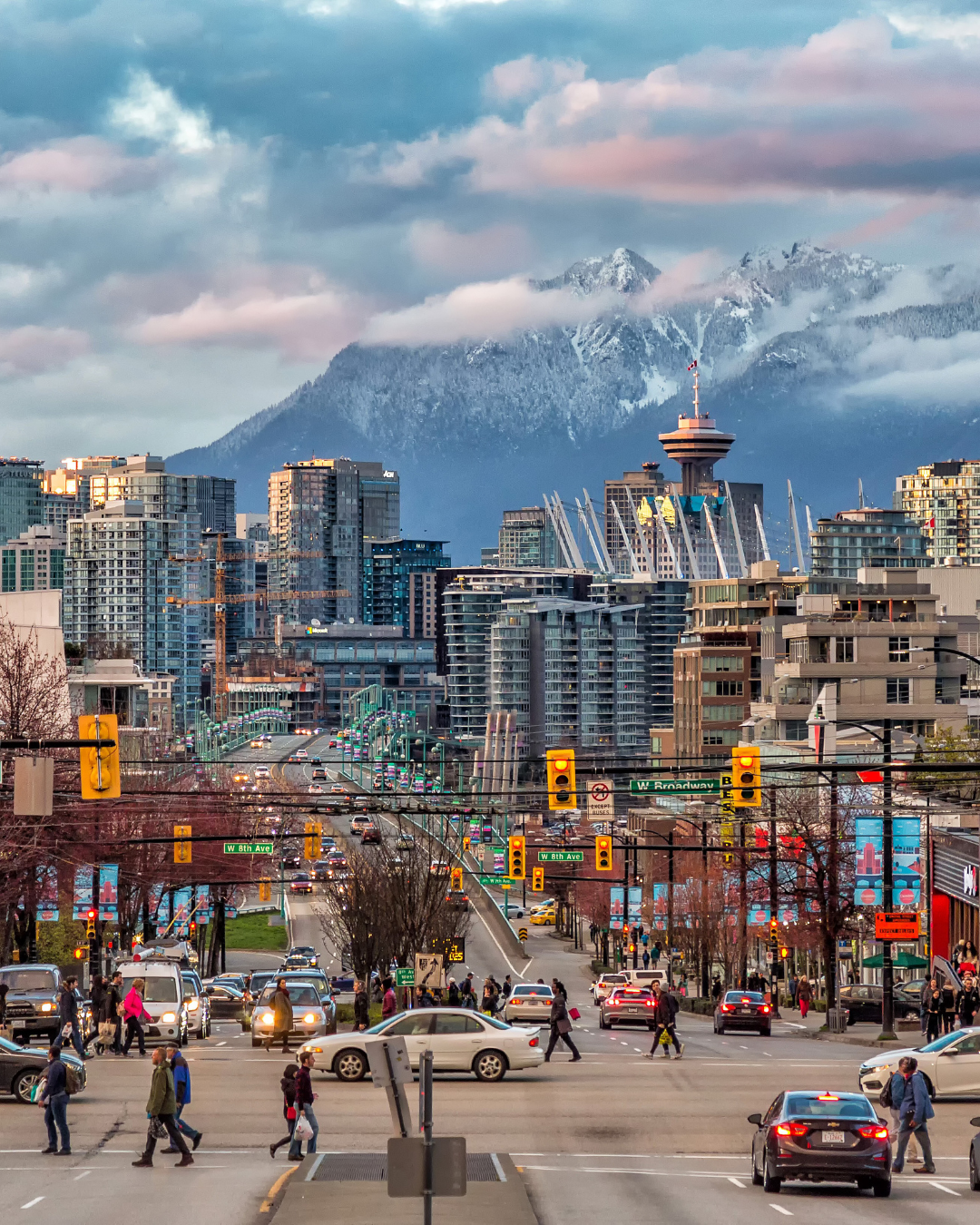 Bustling downtown Vancouver with people crossing the street, modern cityscape, and snow-covered mountains in the background.