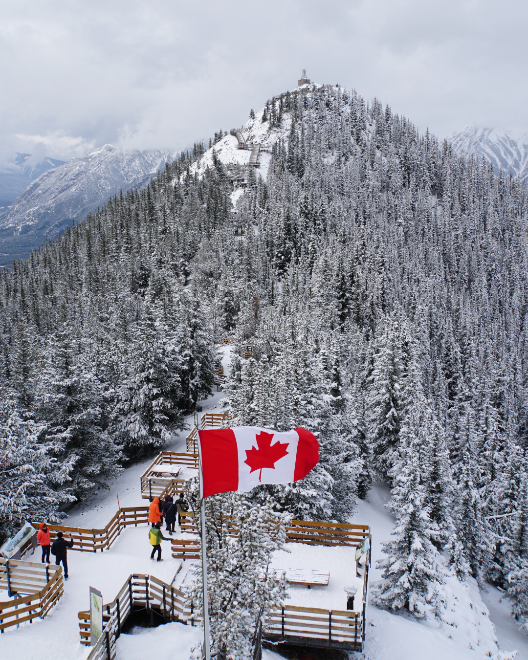 Snow-covered wooden pathways in Banff, Alberta, with people walking through a wintry forest and a Canadian flag flying in the foreground.
