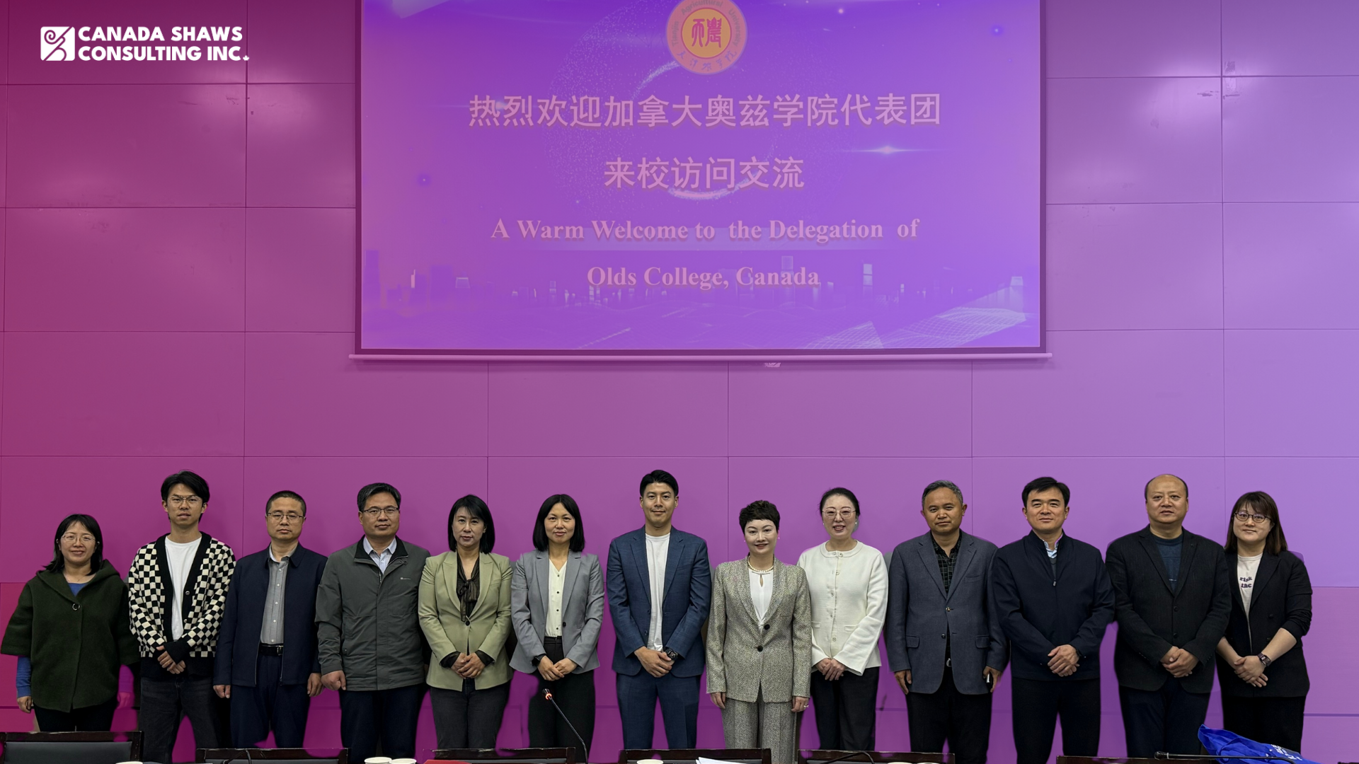 Group photo of representatives from Canada Shaws, Olds College, and Tianjin Agricultural University standing in front of a purple wall with a projection screen displaying a warm welcome message for the Olds College delegation. The welcome screen features both English and Chinese text.