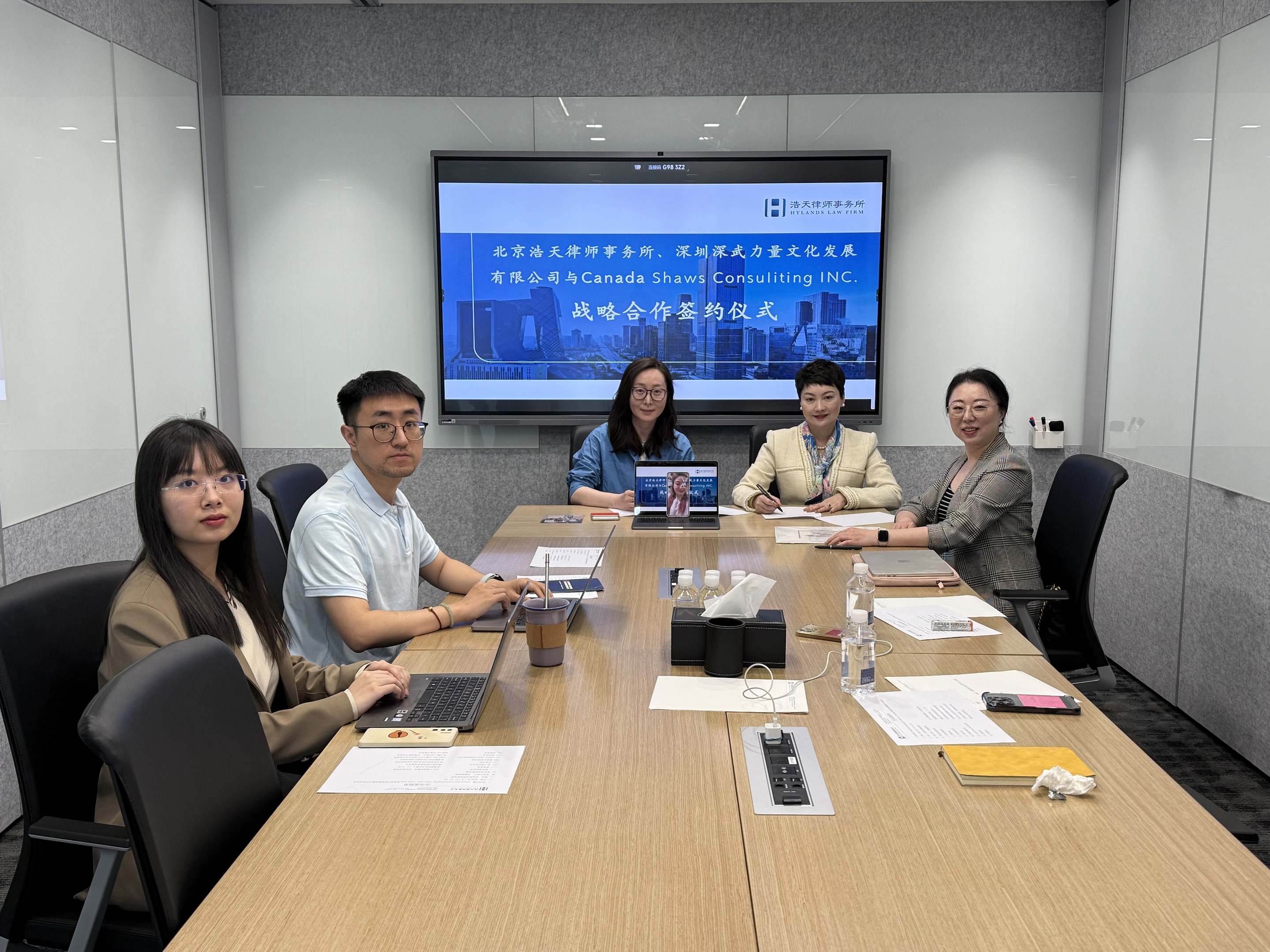 Delegates from Canada Shaws and Hylands Law Firm sit around a conference table, moments before or after signing their strategic partnership agreement. A large screen behind them displays the event title in Chinese, with the names of both firms. Laptops, documents, and refreshments are visible on the table, emphasizing a formal yet collaborative meeting atmosphere.