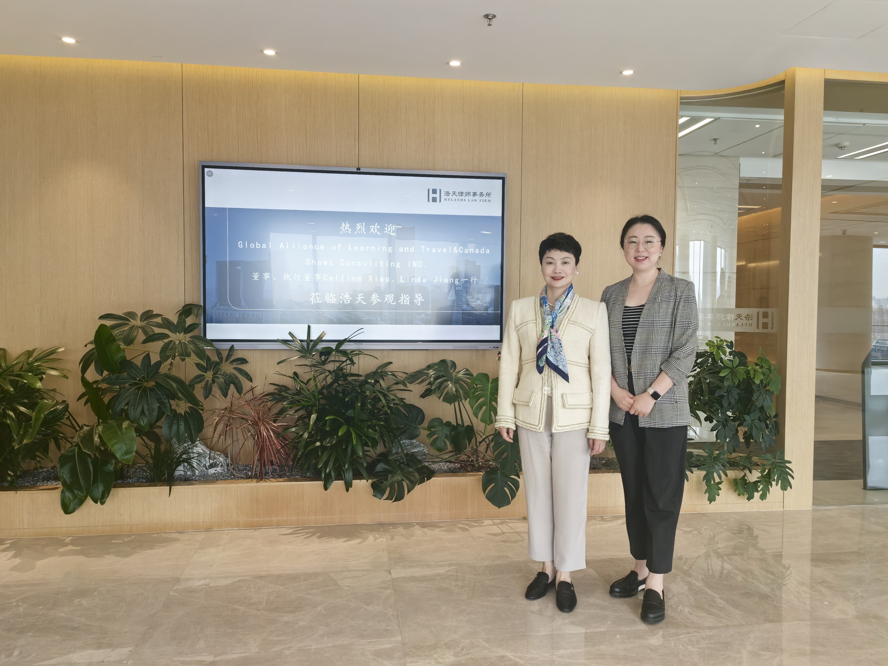 Two women pose together in front of a large digital welcome screen at Hylands Law Firm’s office. The screen displays a personalized message welcoming representatives from Canada Shaws and their affiliated organizations. They stand amid green indoor plants with modern wood-panel decor surrounding them.